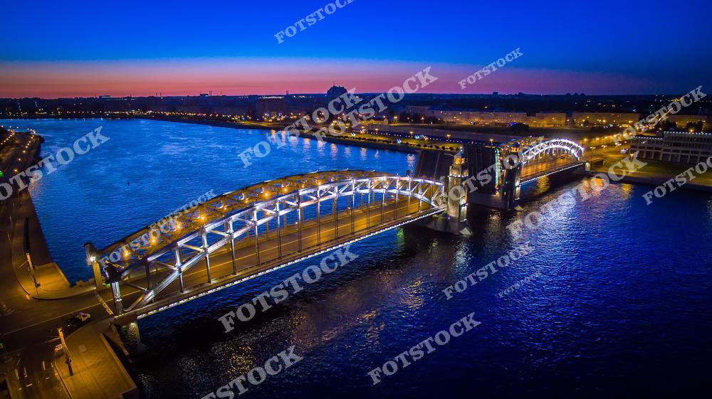 На фотографии изображен Большеохтинский мост в Санкт-Петербурге, освещенный яркими огнями. Мост, перекинутый через реку Неву, выглядит особенно величественно в вечернем свете. Спокойная поверхность воды отражает свет мостовых конструкций, создавая живописный вид. Этот мост, одно из знаковых сооружений Петербурга, передает дух и красоту города ночью.